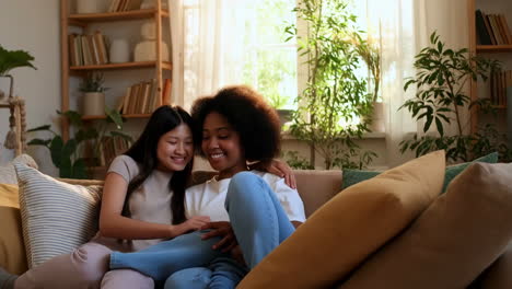 two diverse young women hugging and smiling on a couch at home