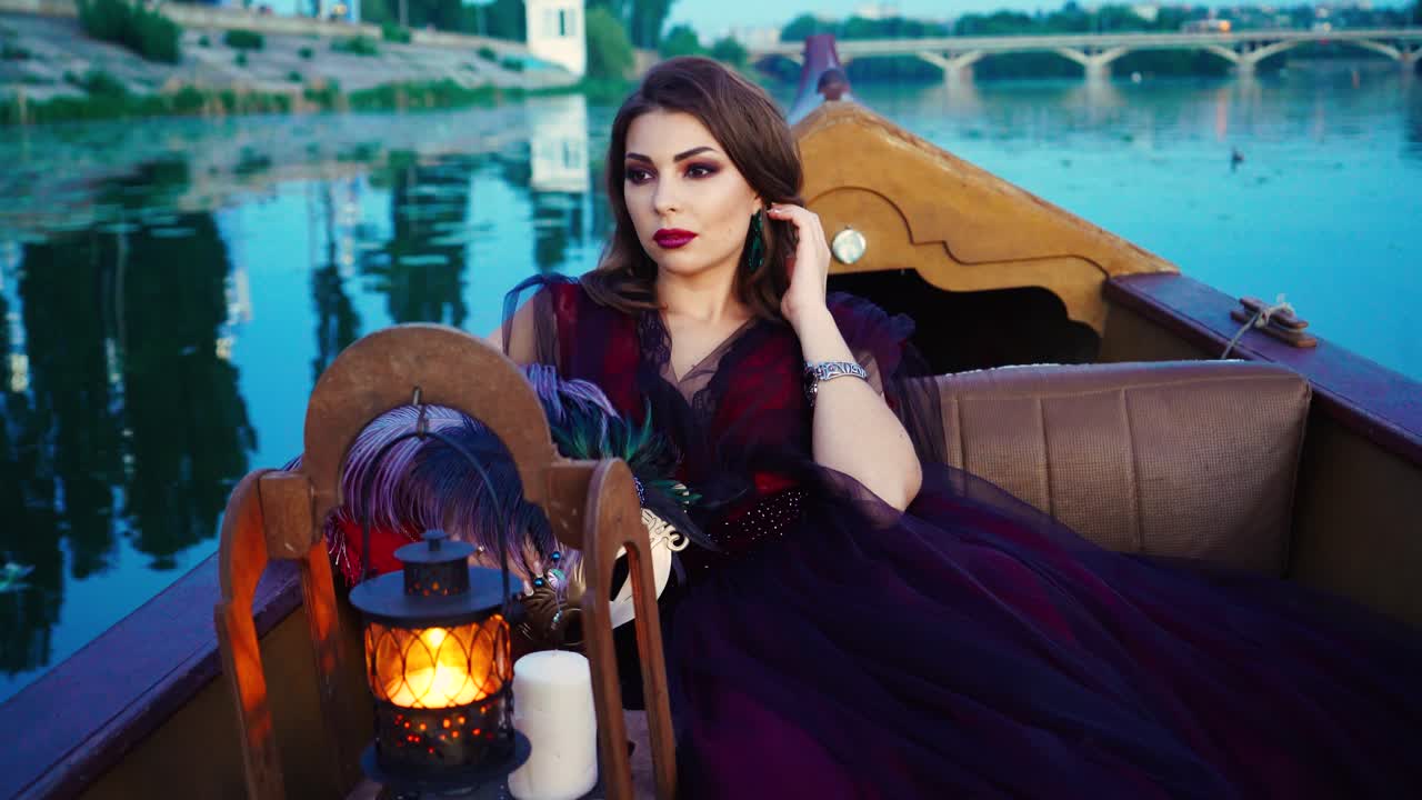 A woman with make-up is sitting in the gondola and holding mask in her hands with a colored feathers near a decorative lantern and white candles on the background of the river in the evening. Carnival in Venice
