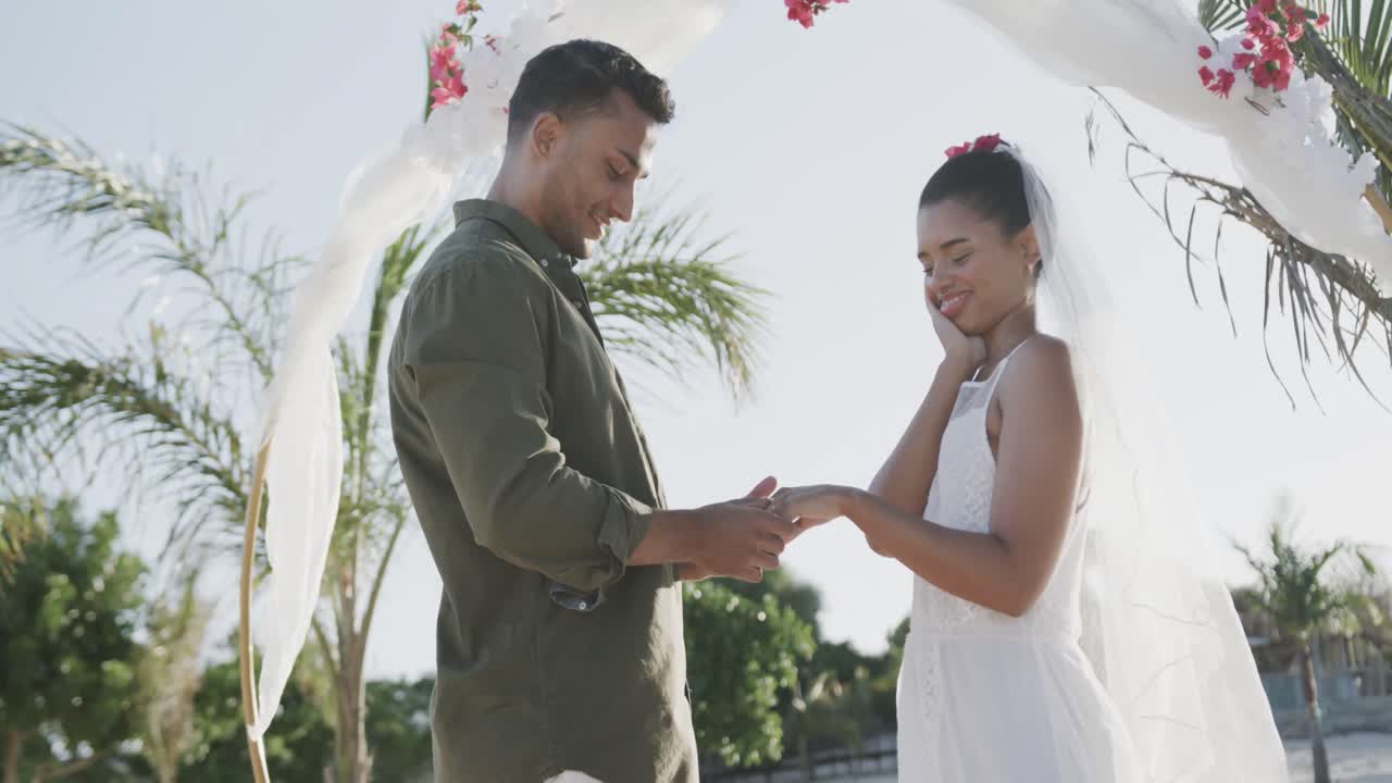Diverse groom putting wedding ring on finger of bride and embracing at beach wedding, in slow motion