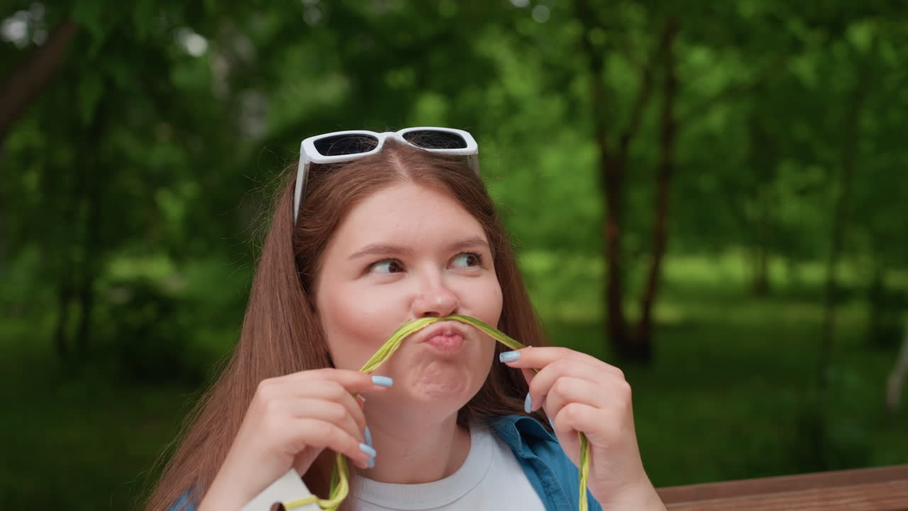 Close up of young woman sitting outdoors holding colorful thread across face pretending mustache while making funny playful expression, laughing cheerfully in green park