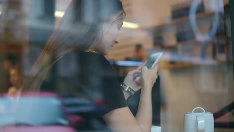 Vista-Desde-El-Exterior-De-Una-Joven-Concentrada-Con-El-Pelo-Largo-Enviando-Mensajes-Usando-Su-Teléfono-Móvil-En-La-Toma-De-Café.-Taza-Blanca