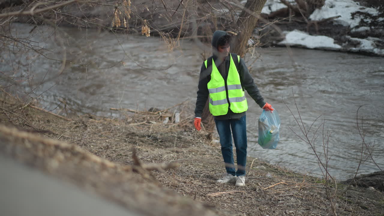 Volunteer wearing reflective vest and gloves collects litter along riverbank, placing trash into blue plastic bag during community cleanup to protect environment and improve natural habitat