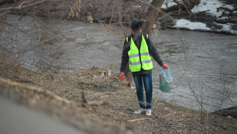 volunteer wearing reflective vest and gloves collects litter along riverbank, placing trash into blue plastic bag during community cleanup to protect environment and improve natural habitat