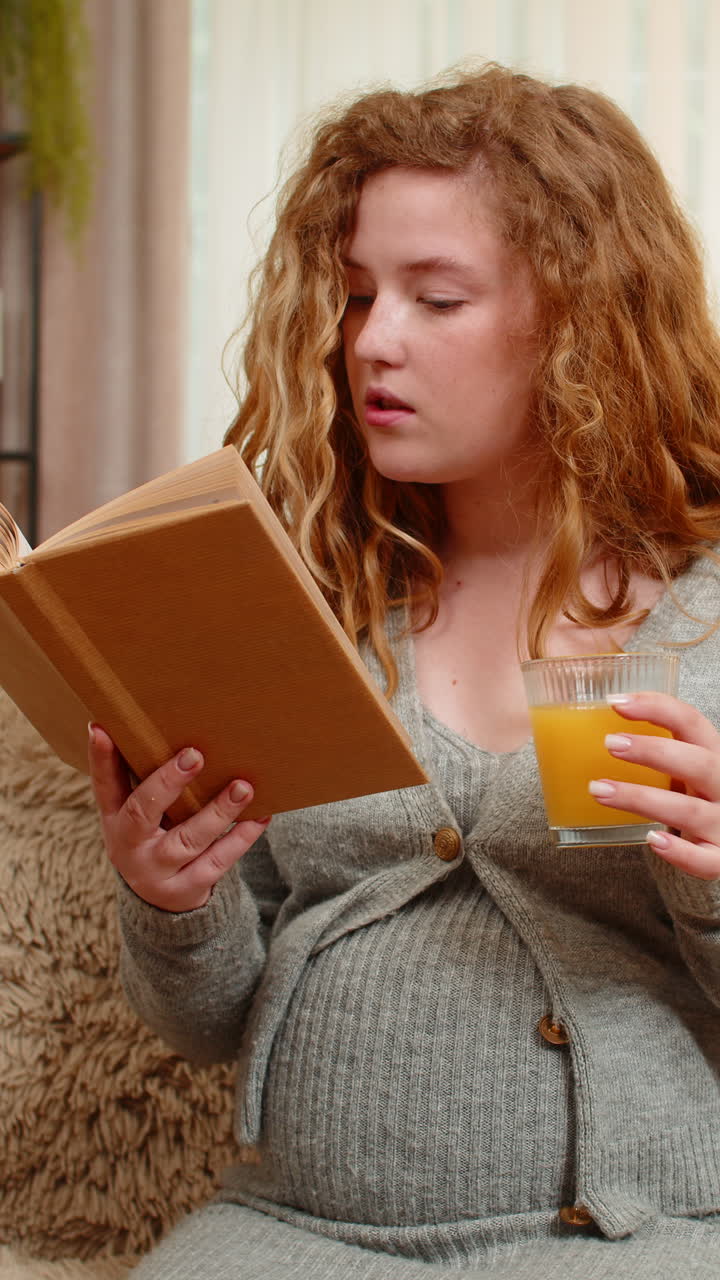 Pregnant young woman drinking orange juice while reading pregnancy book at home calm relaxed mood