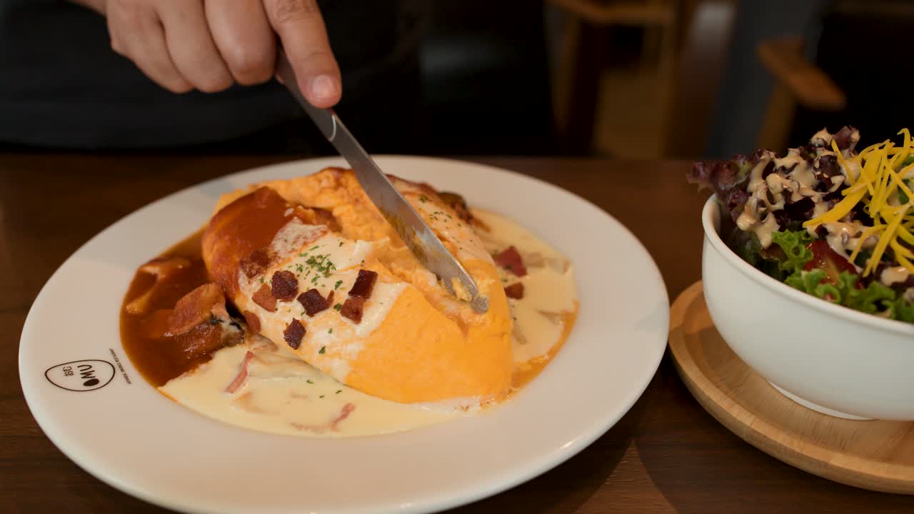 Hand slices omelette rice with creamy sauce, salad on side, warm restaurant lighting, steady close-up