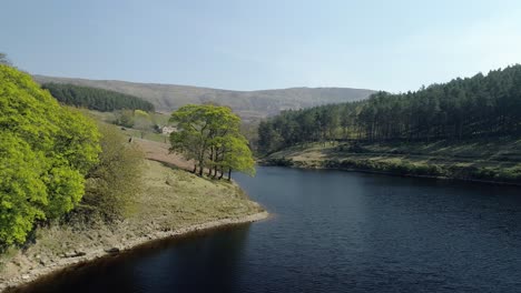 Aerial-shot-panning-right-over-Kinder-Reservoir-water-close-to-trees-revealing-a-National-Trust-estate-in-the-distance-in-the-Kinder-Scout-Valley