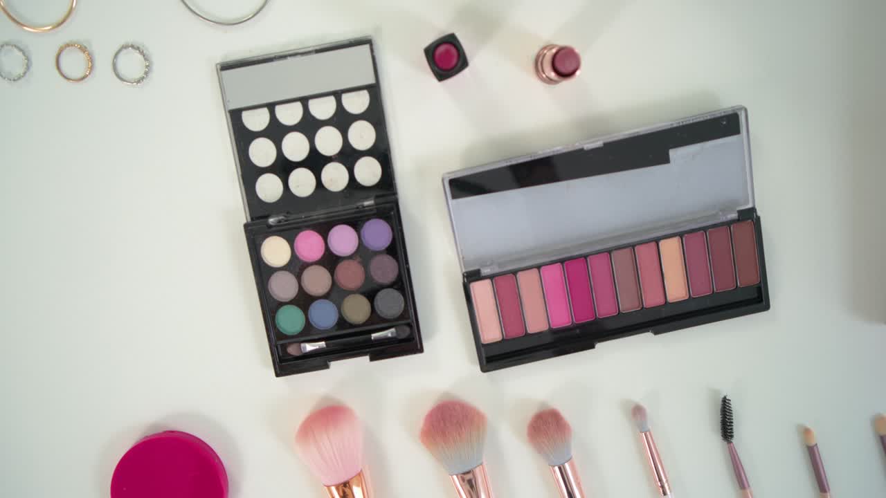 Camera pulling back panning over white table revealing brushes palettes lipsticks rings for display