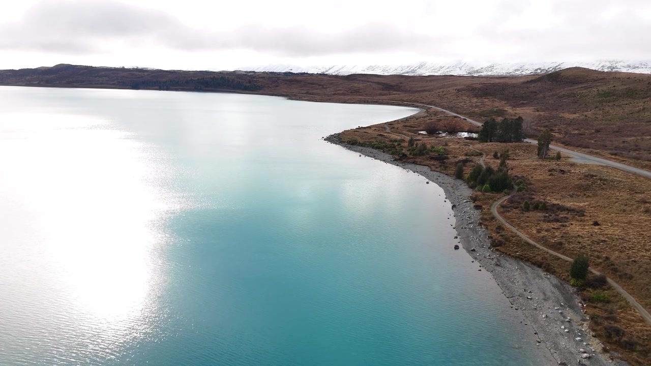 Filmagem aérea de drone para frente ao longo da margem do Lago Pukaki, exibindo águas glaciais de um turquesa vívido, terreno acidentado e paisagens alpinas deslumbrantes na Nova Zelândia.