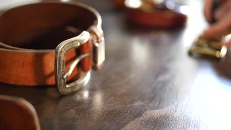 close up on a leather workers workbench with worn leather belts with heavy patina on the buckle as he works on a new belt