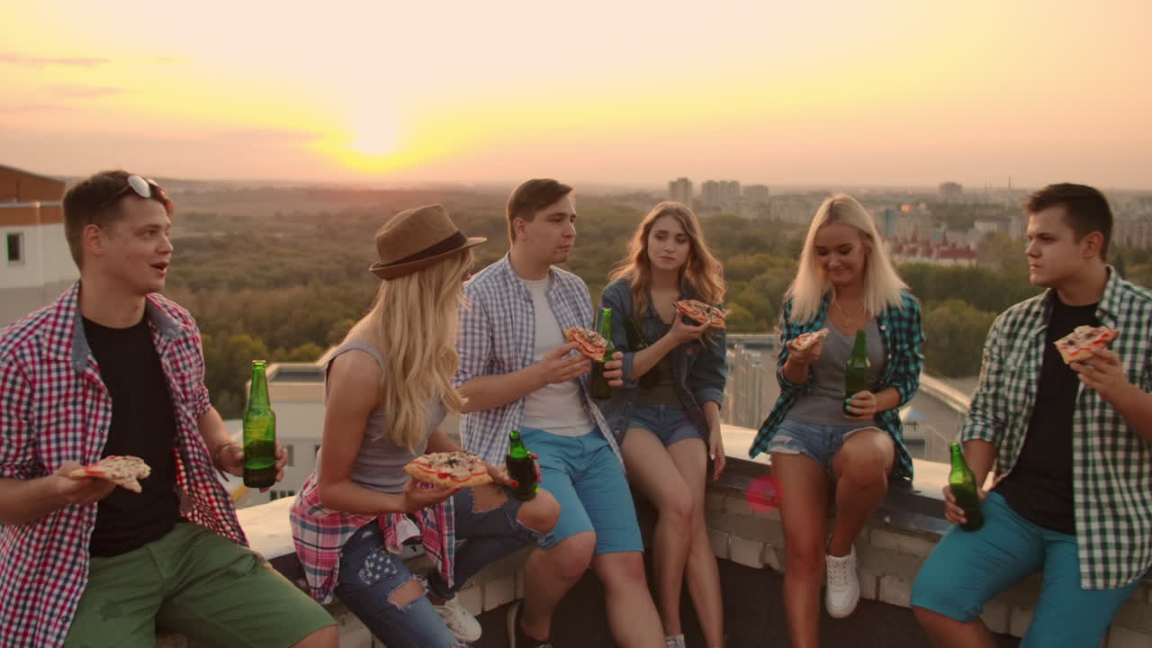 Three girls and three boys in plaid shirts clinks glasses and drinks beer from green bottels on the party with friends on the roof at the sunset.They are sitting together eat hot pizza in summer evening.