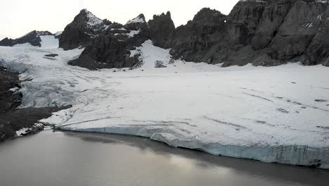 A-spinning-aerial-flyover-over-the-glacial-lake-of-Claridenfirn-glacier-in-Uri,-Swizerland-at-dusk-with-a-view-of-the-glowing-sky-behind-the-alpine-peaks-and-floating-icebergs-in-the-water