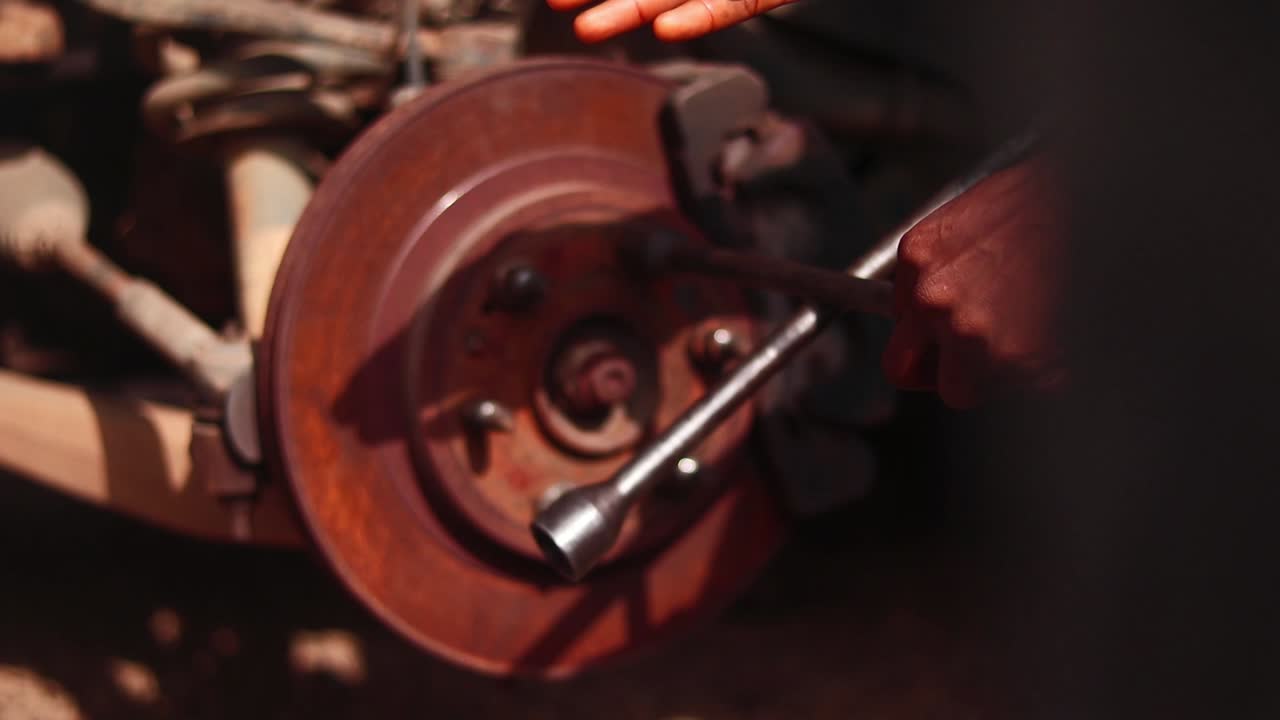Fixed camera close-up of a mechanic’s hand using a tool on a rusted car brake in Nigeria.