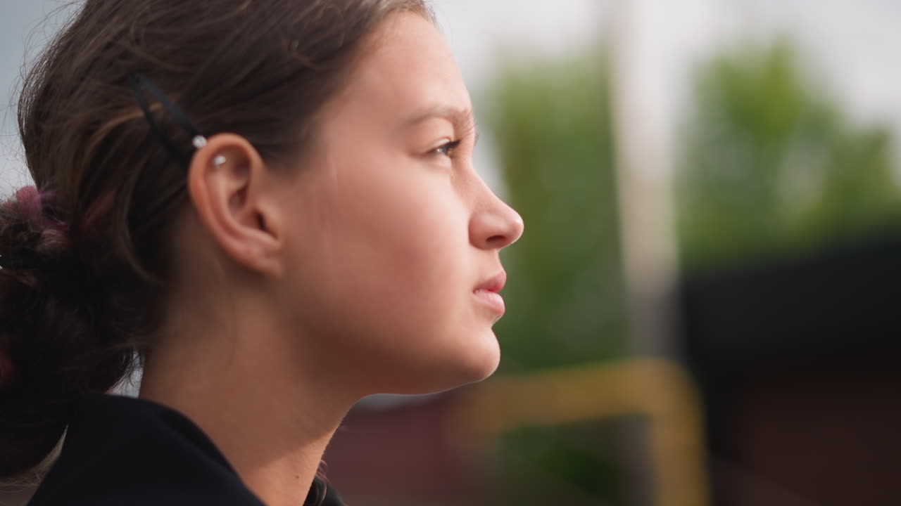 Profilo di adolescente sotto una pioggia leggera, occhi chiusi ed espressione calma, gocce di pioggia nei capelli, sfondo suburbano all'aperto con vegetazione e cielo coperto, umore riflessivo e brezza leggera, riflessi nelle pozzanghere nelle vicinanze.