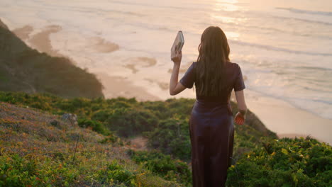 Back-view-woman-walking-down-green-grass-hill-in-front-sunset-seascape.