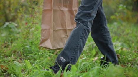 leg view of boy wearing dark jeans and black sneakers walking through vibrant greenery, holding brown paper bag, surrounded by lush plants and foliage in outdoor natural setting