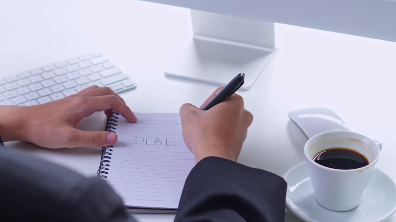 Businessman in suit writes the word "DEAL" in spiral notebook on white desk with coffee and computer