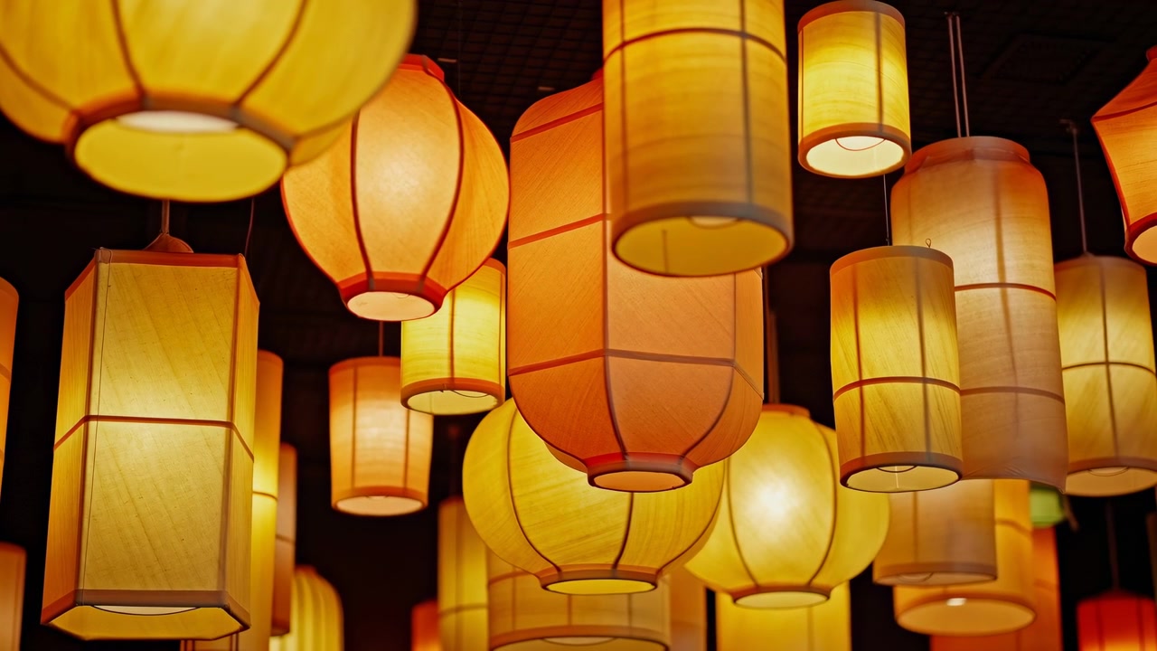Numerous hanging paper and fabric lanterns glow above dark interior space, shifting subtly in parallax while suspended from ceiling grid, creating warm decorative atmosphere and depth