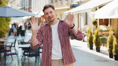 young man smiling friendly at camera waving hands hello hi greeting or goodbye in city street