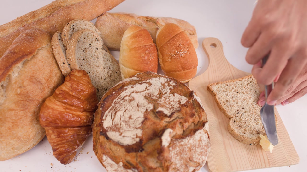 Primo piano di mani che spalmano burro su pane ai cereali, accanto a pane a lievitazione naturale, croissant e baguette