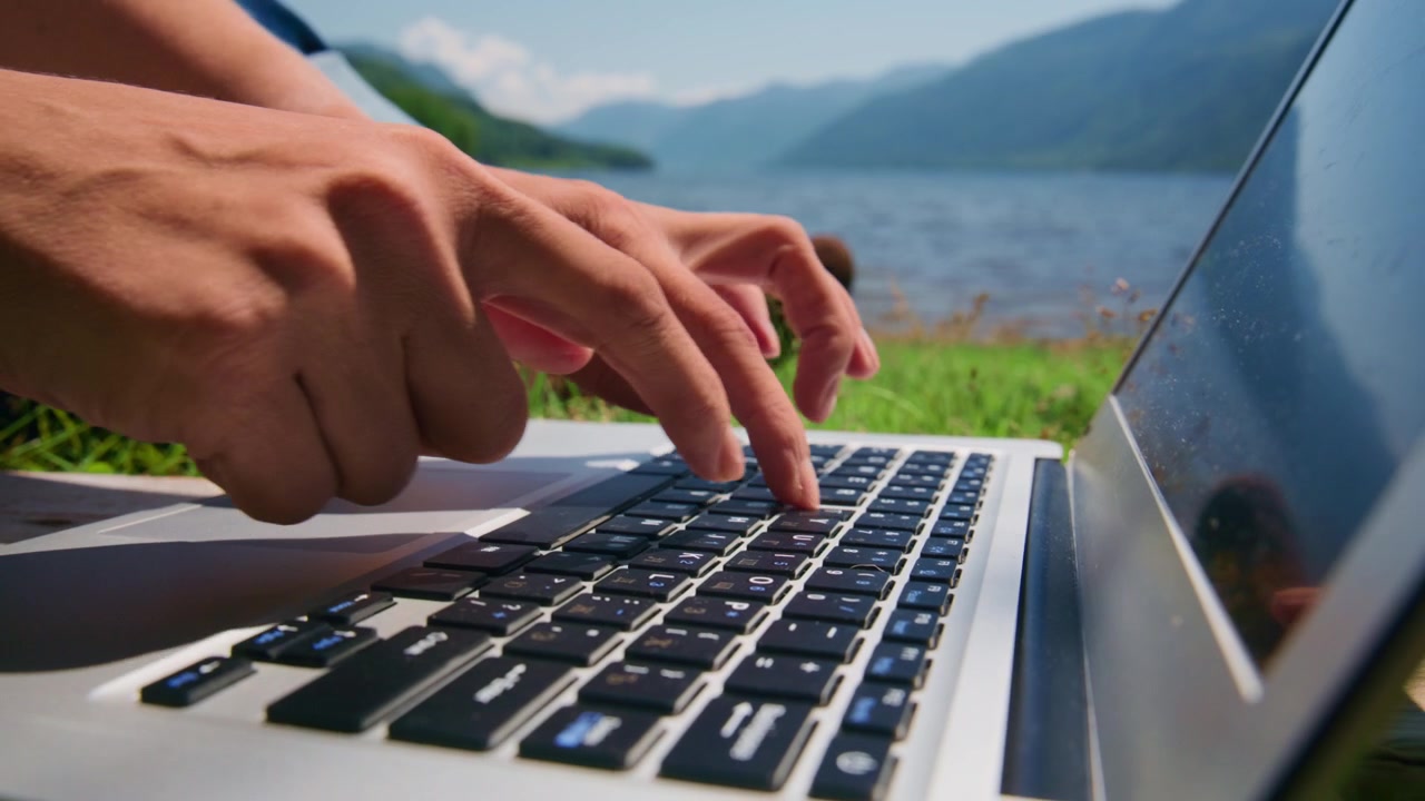 Hands type on laptop keyboard, outdoors near lake. Mountains and green grass behind. Person focused, calm, working remotely. Perfect for remote work, travel, or nature-themed content