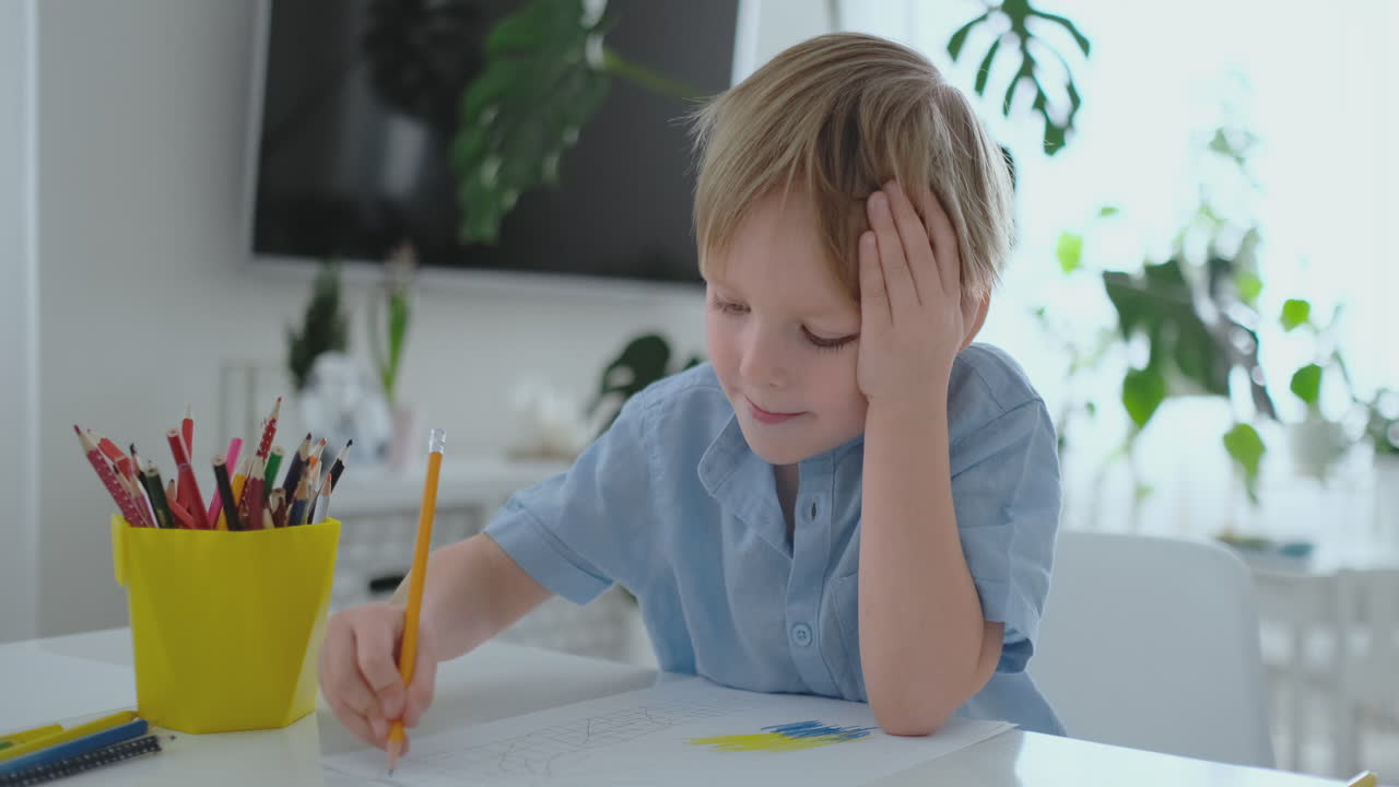 A boy in a blue t-shirt sitting in the kitchen at the table draws a pencil doing homework preschool training