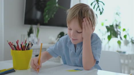 Un-Niño-Con-Una-Camiseta-Azul-Sentado-En-La-Cocina-En-La-Mesa-Dibuja-Un-Lápiz-Haciendo-La-Tarea-De-Entrenamiento-Preescolar