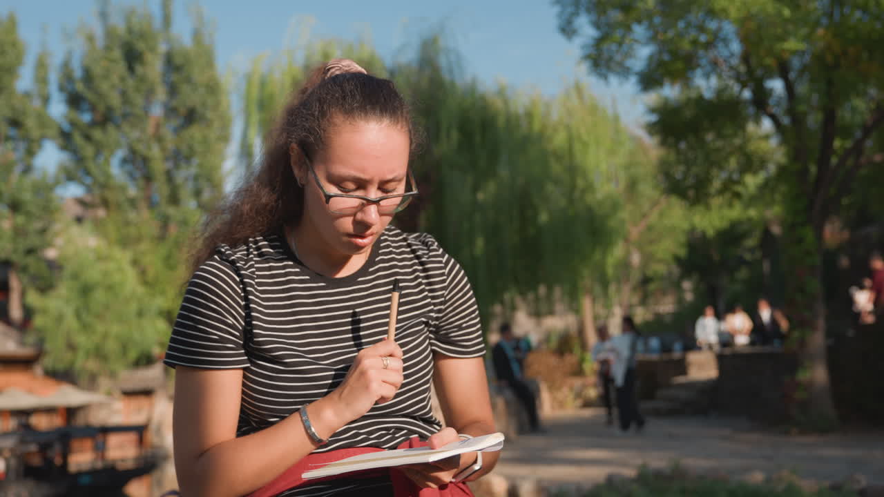 Giovane donna con i capelli ricci prende appunti all'aperto. Studentessa con zaino studia in un ambiente universitario soleggiato. Studentessa casual con occhiali e capelli ricci si concentra sul prendere appunti all'aperto.