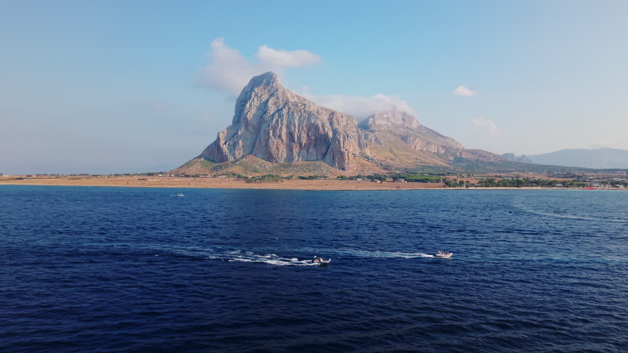 Drone shot over the sea orbiting boats with a towering coastal rock and wild beach in Sicily