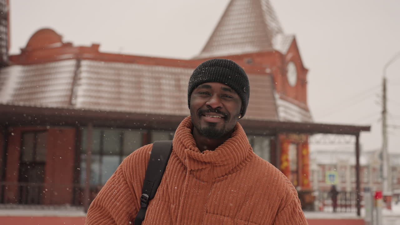 Uomo di colore con cappotto arancione che sorride, zaino in spalla, berretto e occhiali da sole, neve che cade fuori dalla stazione di mattoni catturando un'espressione calda e spontanea, atmosfera di pendolarismo invernale con texture accoglienti e delicate