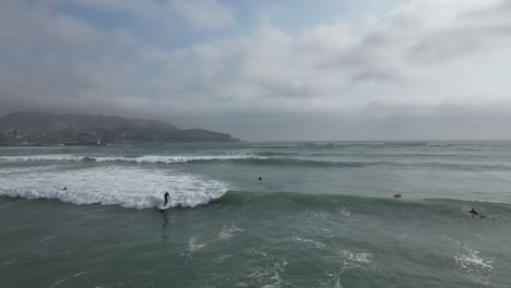 Toma-Aérea-Baja-Del-Hermoso-Mar-Turquesa-En-Miraflores-Mientras-Un-Surfista-Monta-Su-Tabla-De-Surf-En-Las-Tranquilas-Olas-Y-Otras-Personas-Nadan-En-El-Agua