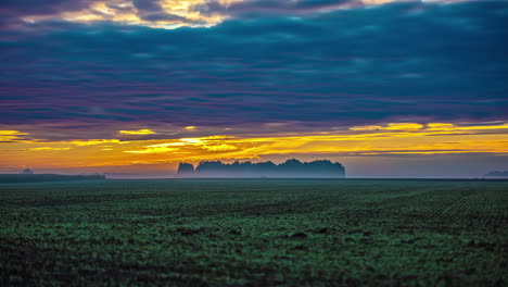 Time-lapse-shot-of-yellow-sunrise-behind-flying-dark-clouds-and-fog-over-farm-field