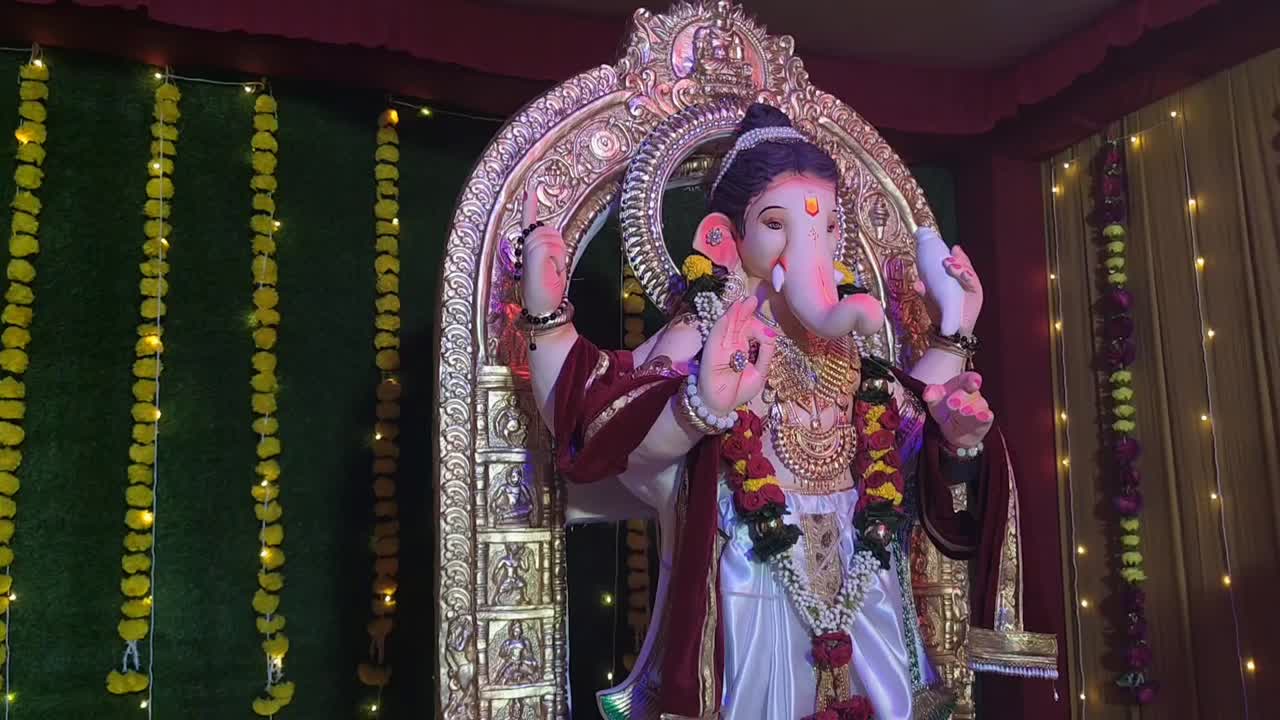 Cross angled view of beautifully adorned Lord Ganesha statue in a pandal, illuminated by focused lighting with vibrant grass and floral backdrop.