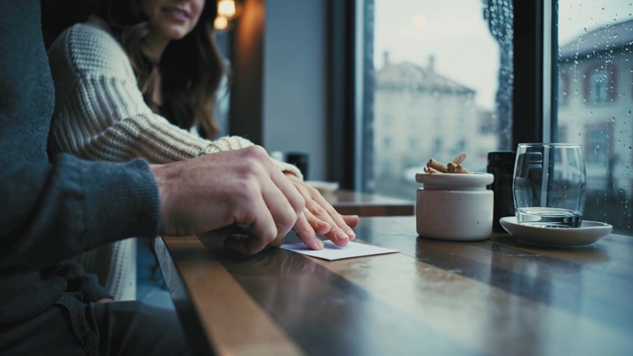 Uomo che batte le dita con maglione grigio, donna con maglione bianco che guida e passa una busta al tavolo del caffè