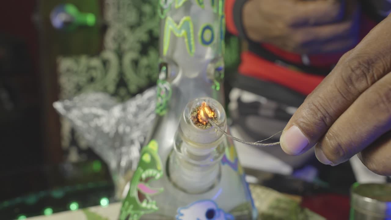 un hombre negro usando una mecha para encender un bong de agua de marihuana lleno de hielo y se inclina para verlo exhalar el humo