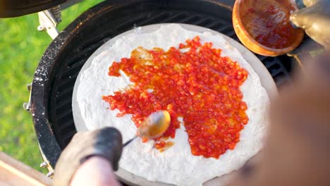 chef topping tomato sauce with spoon on pizza grilling outdoors, close-up