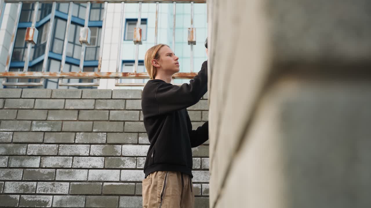 Weißer Mann untersucht eine Ziegelmauer, hält Kamera, blickt nach oben, urbane Treppe und Betonfassade, Cargohose und schwarzer Pullover, nachdenkliche Atmosphäre und authentische Bildgestaltung, testet Winkel und Textur