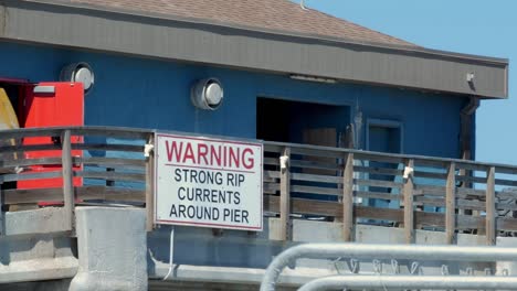 Close-up-of-the-Bob-Hall-Pier-building-in-Corpus-Christi,-Texas-with-a-sign-saying-"Warning:-Strong-Rip-Currents-Around-Pier"-as-a-bulldozer-drives-on-the-pier