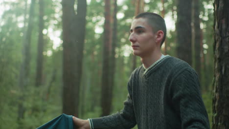close up of boy lifting tent cover on forest ground during camping as soft sunlight filters through trees, smiling slightly while working with someone nearby to prepare campsite beside tree trunk