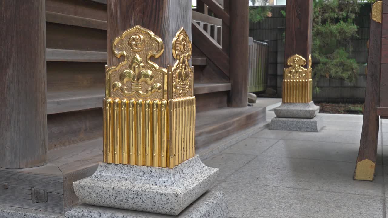 A close-up view of the wooden columns at Kuramae Shrine, adorned with intricate golden decorative metalwork atop a stone base.