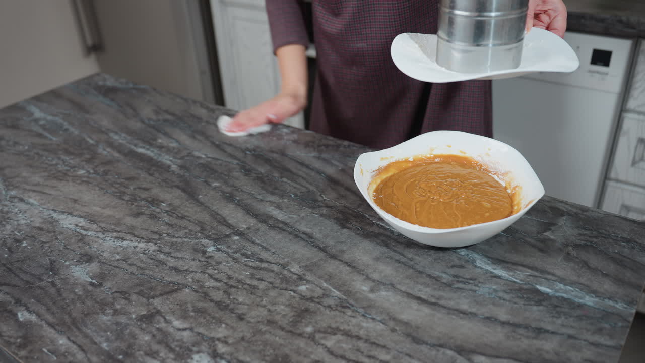 vista parcial de una mujer limpiando la encimera de la cocina con un paño mientras lleva un tamiz en el plato, un cuenco de masa se sienta en la superficie de mármol, destacando la preparación de alimentos y la limpieza en la cocina organizada