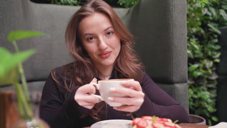 young woman with long brown hair sits comfortably in cafe booth sipping latte from white cup while looking relaxed, table displays waffles topped with strawberries and decorative plant in glass vase