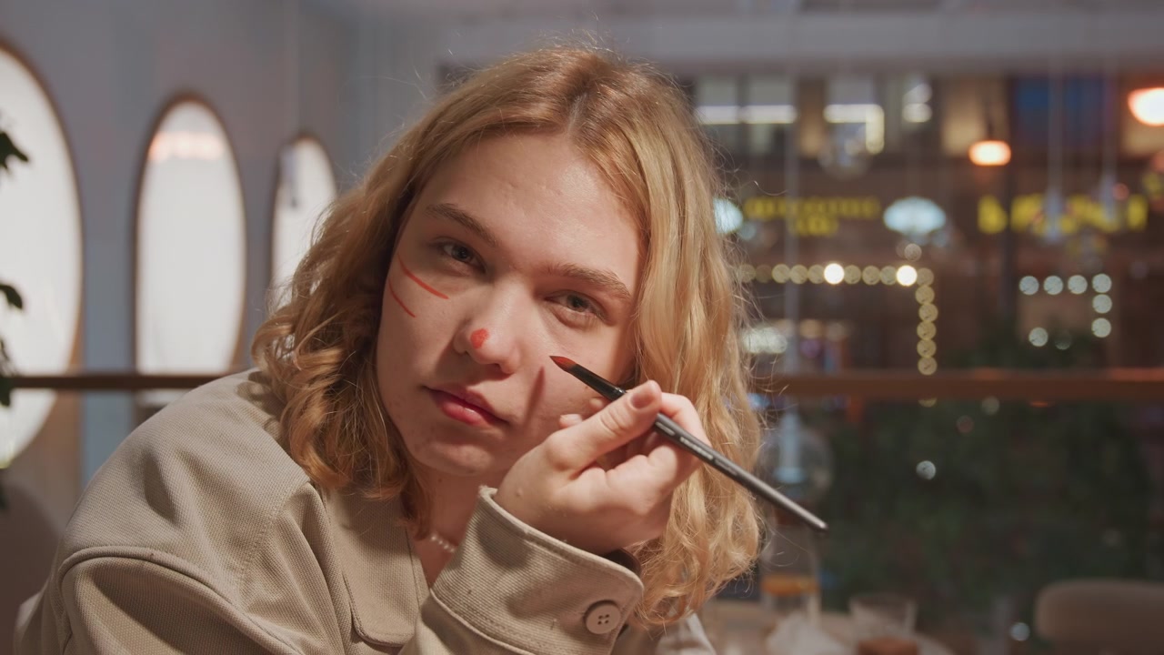 Close up of young woman applying makeup with orange brush in soft lighting. Woman wears beige jacket and pearl necklace seated at what appears to be cafe or restaurant setting visible