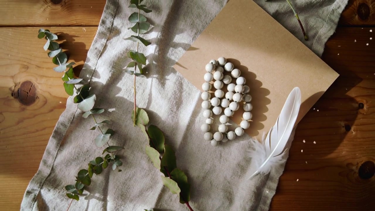 Gliding feather sliding across wood table, pushed by air, with beads and eucalyptus, copy space