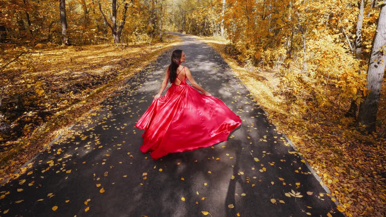 A Glimpse of Autumn Elegance: A Woman in a Flowing Red Dress Walking Down a Leaf-Covered Pathway Surrounded by Golden Foliage and Beautiful Fall Colors