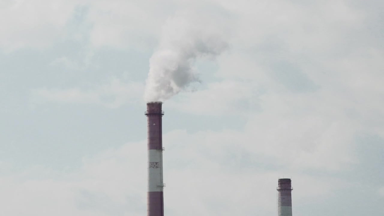 Un'alta ciminiera sulla sinistra emette fumo bianco in un cielo pallido e coperto, con un camino più piccolo visibile in lontananza, creando una chiara scena di intensa attività.