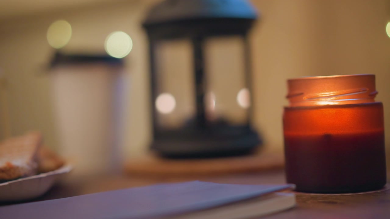 Close up of lit candle in glass jar on wooden table with soft warm light and bokeh background. Notebook lies nearby suggesting quiet workspace or study environment. Candle burns steadily