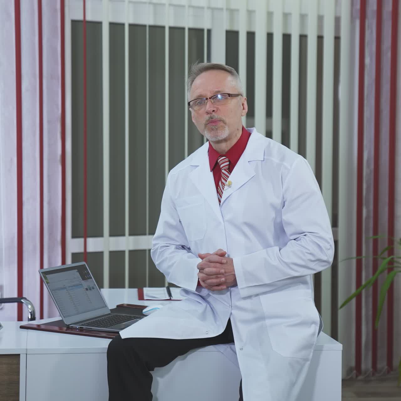 Medical specialist giving online consultation. Friendly senior doctor in white coat and glasses sitting on desk in medical room and explaining something on camera.