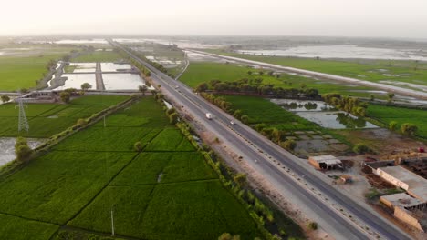 Vista-Aérea-De-La-Carretera-Que-Atraviesa-El-Sindh-Rural-Con-Campos-Anegados-En-Un-Fondo-Lejano