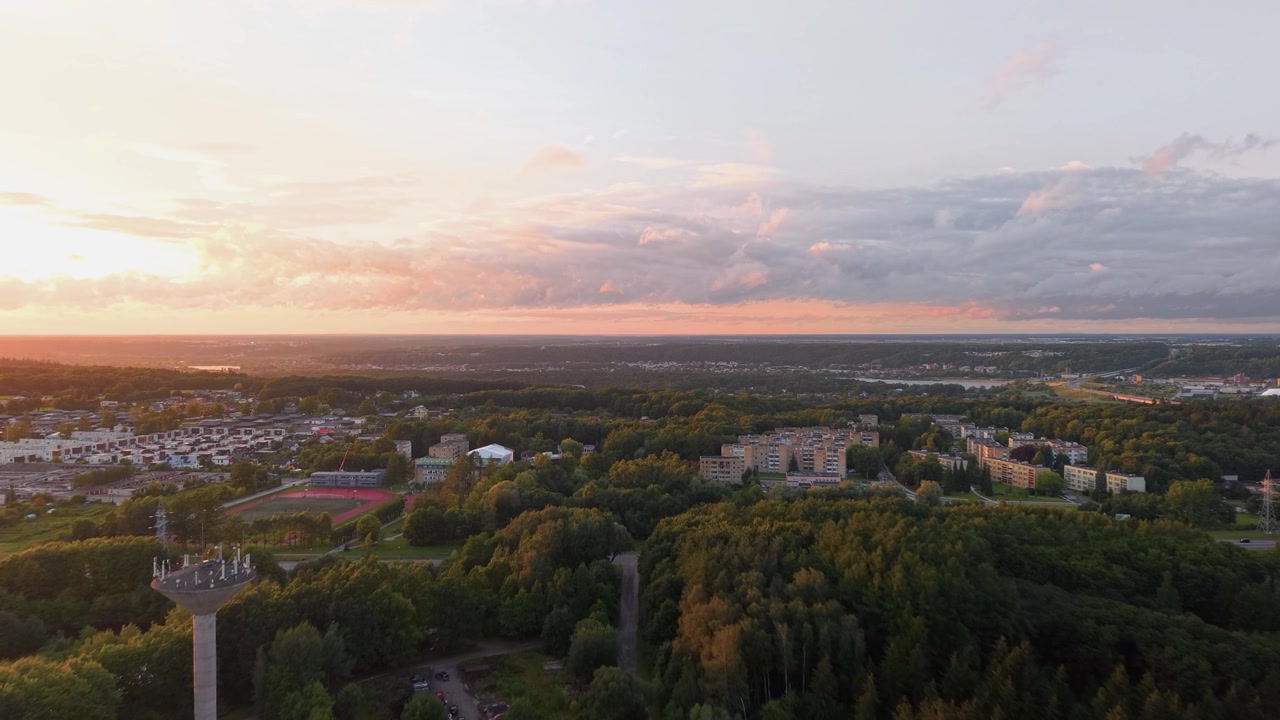 Stunning drone video captures a suburban skyline, lush forest, and distant buildings at sunset. Warm golden light blankets the landscape, dramatic clouds enhance the tranquil, scenic view.