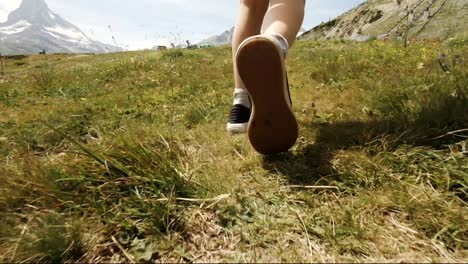 Tiro-De-Carro-Bajo-De-Los-Pies-De-Un-Niño-Caminando-Sobre-La-Hierba-En-El-Paisaje-Montañoso-Cerca-De-Zermatt,-Suiza,-Matterhorn-Parcialmente-Visible-En-El-Fondo